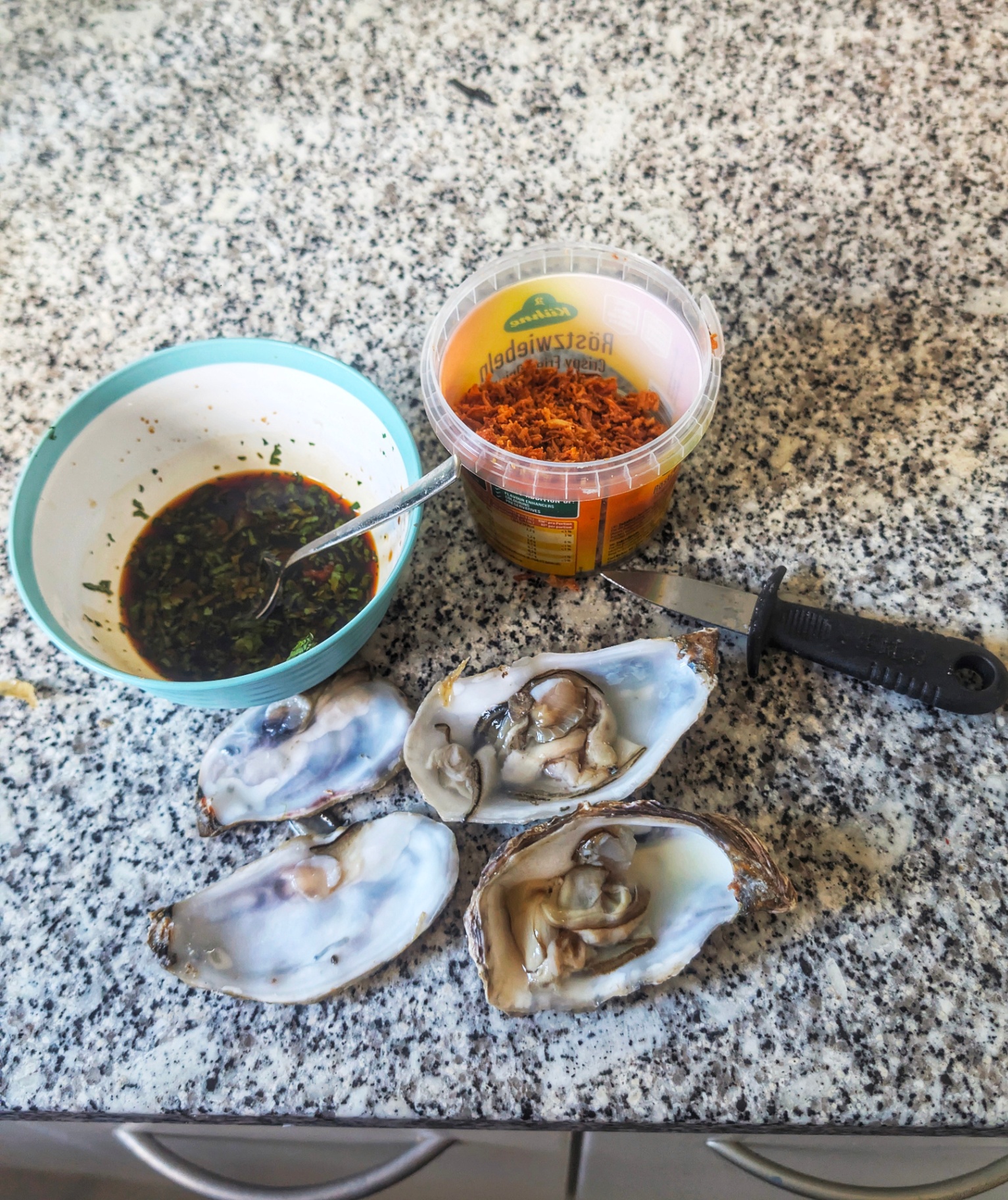Four open oysters on a granite counter, a turquoise bowl of dark chimichurri-style sauce with parsley and a spoon, a tub of crispy fried onions, and a black-handled oyster knife.